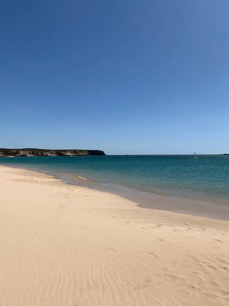 A sandy beach with gentle waves and clear turquoise water under a bright blue sky—one of many tranquil spots among the things to do in Sagres. In the distance, low rocky cliffs border the coastline. The beach appears calm and empty.