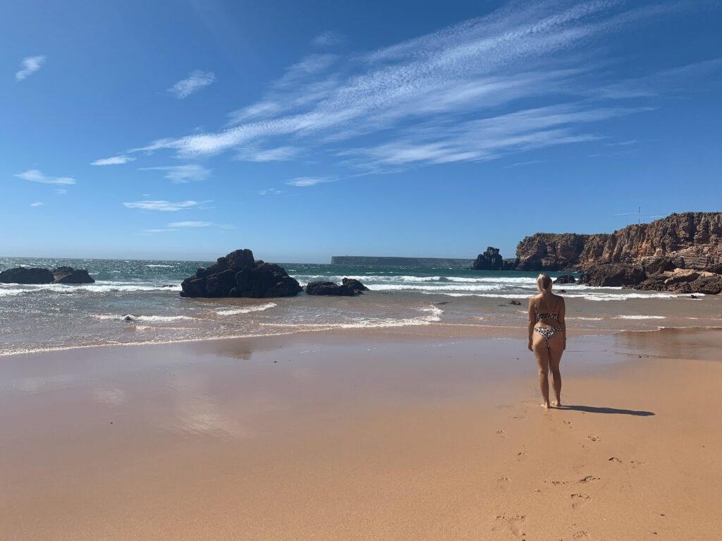 A woman in a bikini walks along a sandy beach toward the ocean, surrounded by waves, rocky formations, and cliffs—one of the many stunning things to do in Sagres under a bright blue sky with wispy clouds.