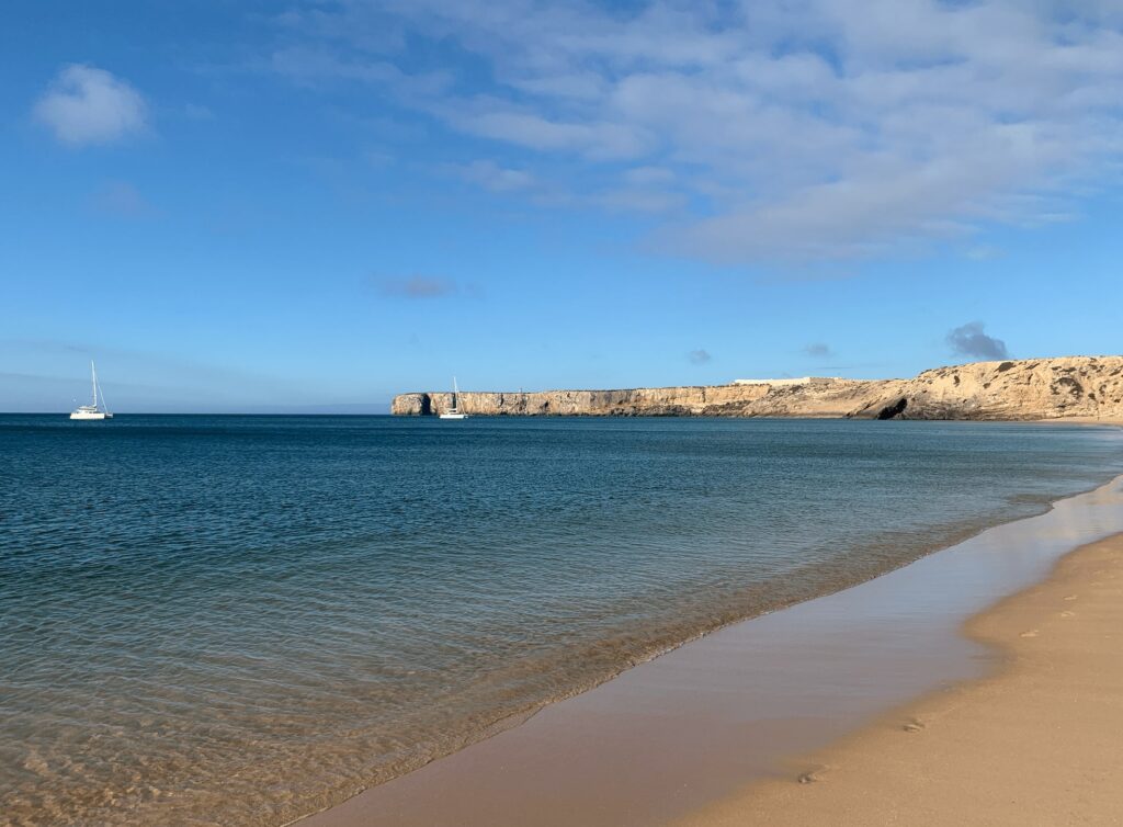 A sandy beach with gentle waves, a calm blue sea, two sailboats gliding by, and rocky cliffs in the distance under a bright blue sky—perfect for anyone exploring things to do in Sagres.