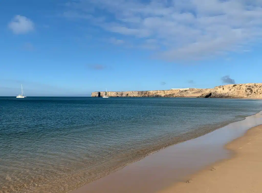 A sandy beach with gentle waves, a calm blue sea, two sailboats gliding by, and rocky cliffs in the distance under a bright blue sky—perfect for anyone exploring things to do in Sagres.