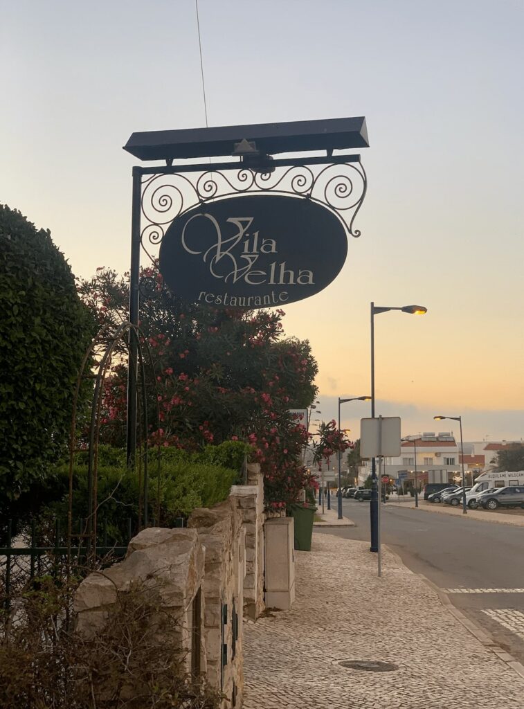 A black sign for "Vila Velha restaurante" hangs above a cobblestone sidewalk lined with bushes and flowering plants, with street lamps and buildings visible at sunset—perfect for those seeking things to do in Sagres.