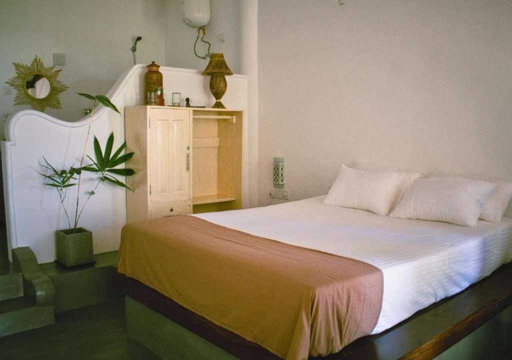 A cozy bedroom reminiscent of charming hotels in Hiriketiya features a neatly made bed with white pillows and a brown throw. A minimalistic wooden cabinet with decorative items and a potted plant adds charm, while soft lighting and a sun-shaped mirror enhance the room's warmth and comfort.