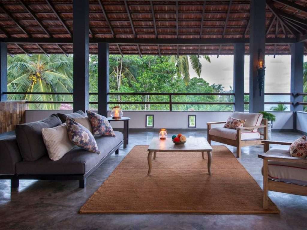  Open-air living space boasts a rustic roof and a gray sofa adorned with floral pillows. Two armchairs flank a wooden coffee table on a textured rug. Tropical trees sway beyond the railing, as candles and fruit add to the serene ambiance.