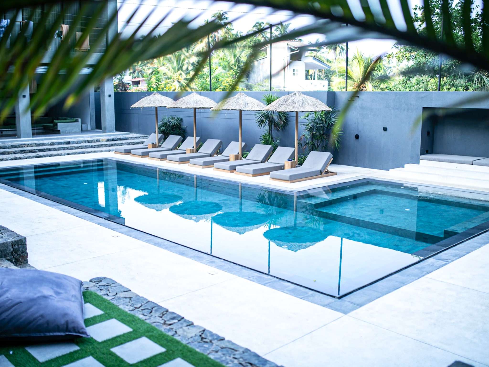 A modern outdoor pool area reminiscent of the serene hotels in Hiriketiya, featuring lounge chairs and straw umbrellas lining one side. Surrounded by lush greenery, the clear water reflects the chairs and umbrellas, while a cushion and patterned mat add charm to the foreground.