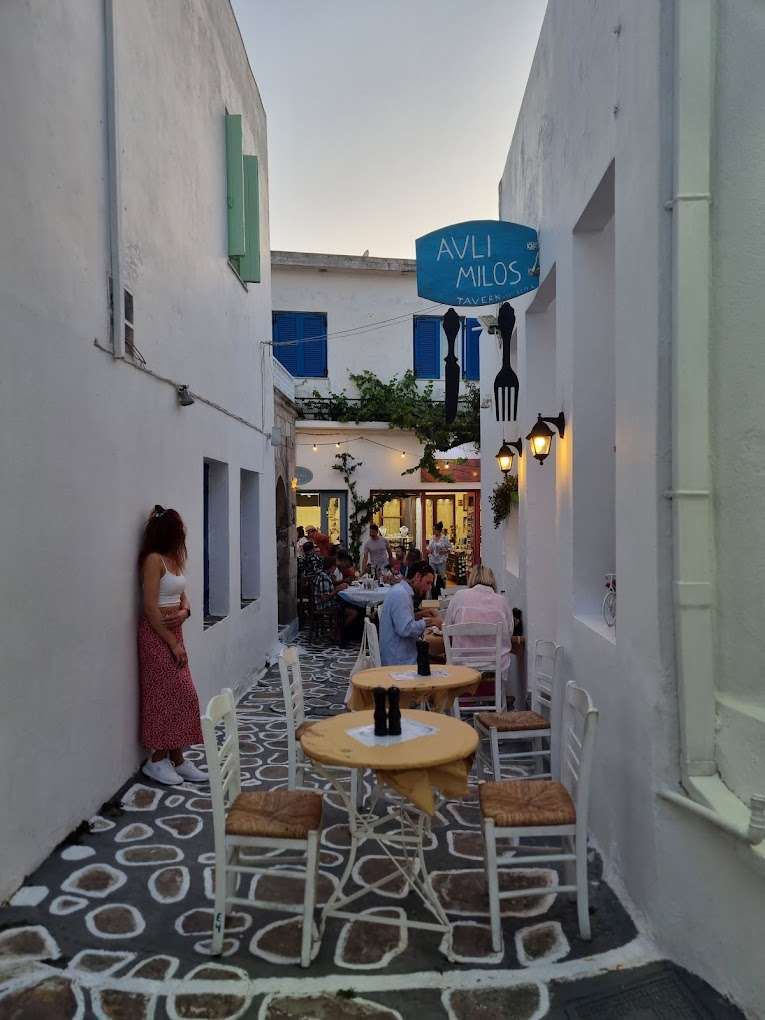 A narrow alley with whitewashed walls leads to a cozy tavern with outdoor seating. People are dining, and a woman stands by the wall. The tavern sign reads "Avli Milos," one of the charming restaurants in Milos. Blue shutters and a warm atmosphere enhance the scene.