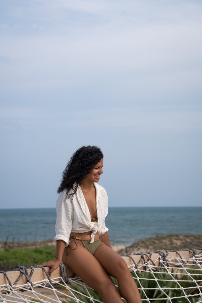 A woman with curly hair, wearing a white shirt and bikini bottoms, sits and smiles on a rope hammock outdoors, with the ocean and blue sky in the backgroundโan image reflecting the freedom often sought by those asking, "Is Sri Lanka safe for solo female travellers?.