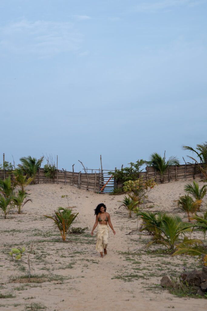 A woman in a light dress walks alone along a sandy path bordered by young palm trees, evoking the question: Is Sri Lanka safe for solo female travellers? A wooden fence and staircase appear in the background under a clear blue sky.
