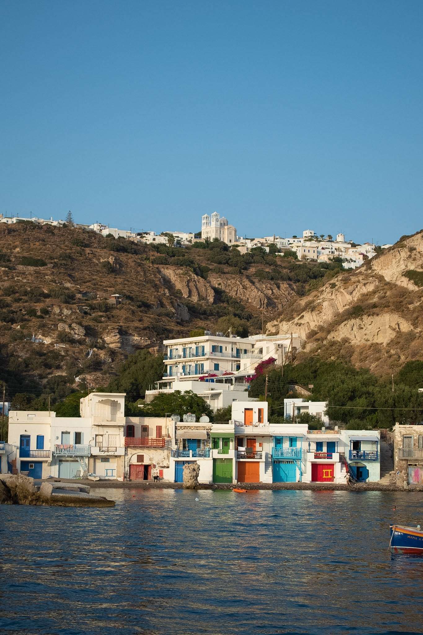 Coastal scene featuring colorful houses along the water's edge, with hills and a small village in the background. The sky is clear and blue, creating a bright and serene atmosphere, reminiscent of the charming ambiance near the restaurants in Milos.