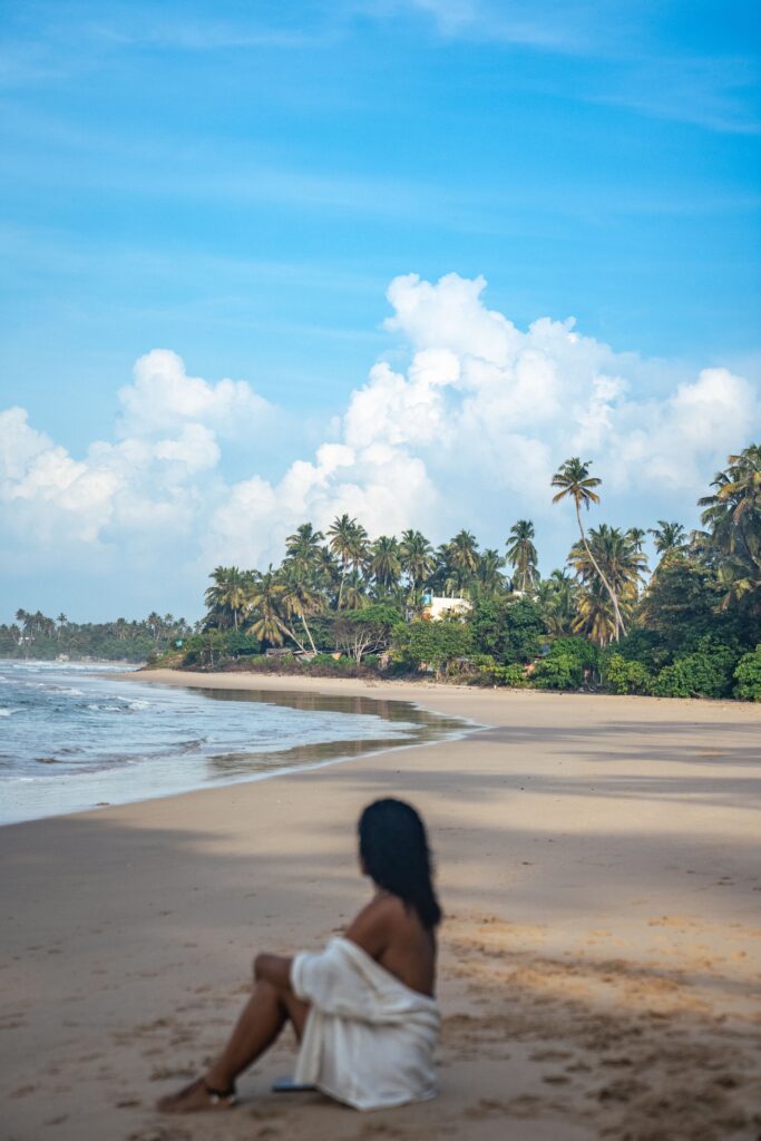 A person sits on a sandy beach wrapped in a white cloth, looking toward the ocean. Palm trees and lush greenery line the shore under a bright blue skyโan inviting scene that may have you wondering, "Is Sri Lanka safe for solo female travellers?.
