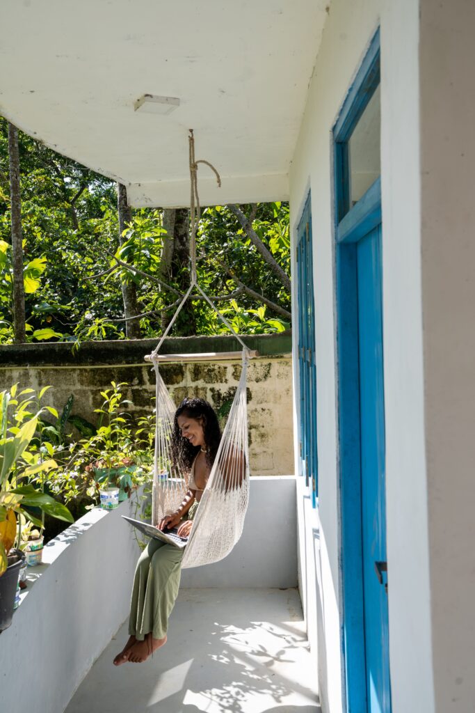 A woman sits in a hanging hammock chair on a sunny balcony, working on a laptop. The bright spaceโwith blue doors, plants, and views of a lush gardenโreflects the relaxed vibe solo female travellers enjoy when asking, "Is Sri Lanka safe for solo female travellers?.
