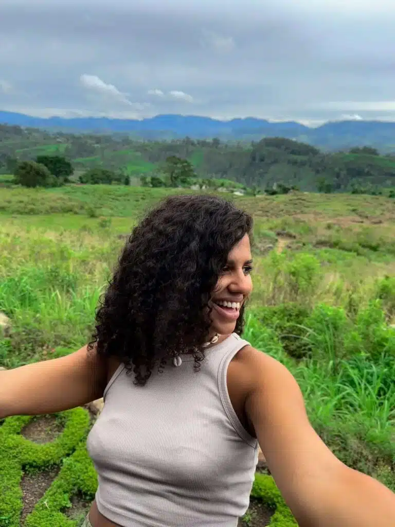 A woman with curly hair and a sleeveless top smiles while taking a selfie outdoors, embodying the spirit of solo female travellers in Sri Lanka, with green fields and distant mountains under a cloudy sky in the background.