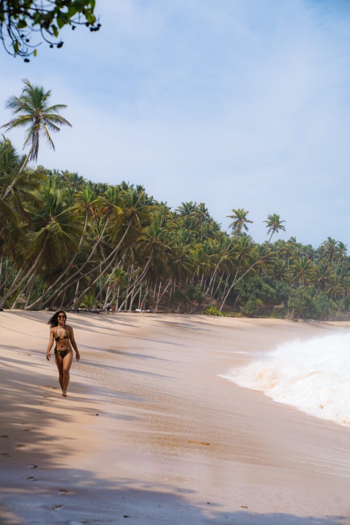 A woman in a bikini walks along a sandy beach lined with tall palm trees, waves crashing onto the shore under a bright blue skyโan inviting scene for those wondering, "Is Sri Lanka safe for solo female travellers?.