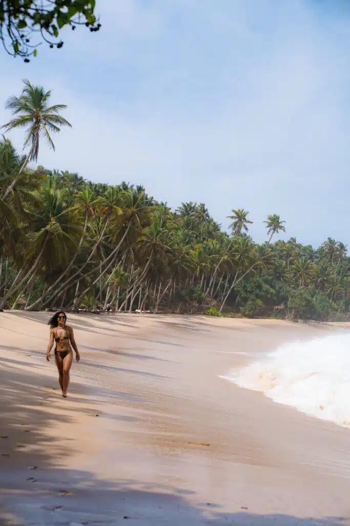 A woman in a bikini walks along a sandy beach lined with tall palm trees, waves crashing onto the shore under a bright blue sky—an inviting scene for those wondering, "Is Sri Lanka safe for solo female travellers?.