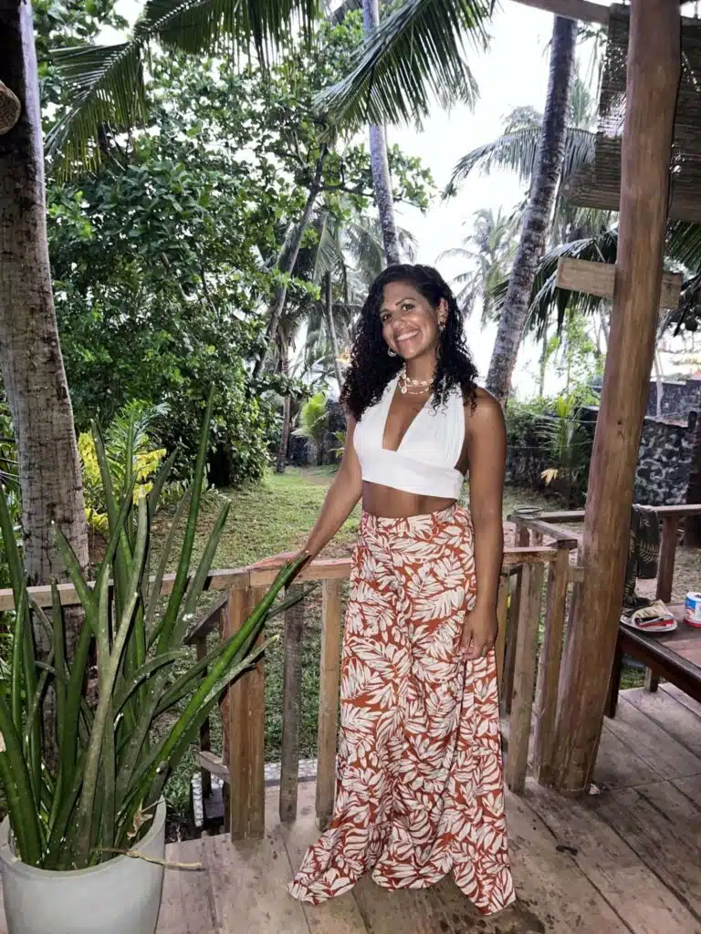 A woman with curly hair, wearing a white crop top and orange leaf-print pants, stands smiling on a wooden porch surrounded by tropical plants and palm trees, reflecting the vibrant beauty that makes many ask: Is Sri Lanka safe for solo female travellers?.