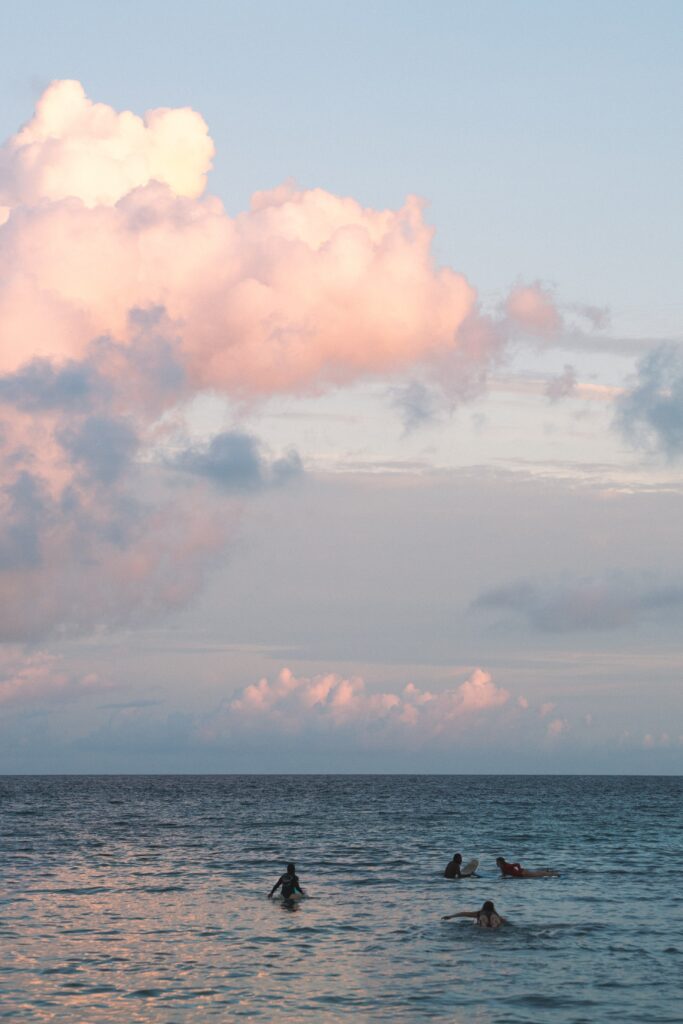 Four people on surfboards float on calm ocean water under a sky with large, soft pink clouds during sunset or sunrise, capturing the tranquil vibe that often makes people wonder: Is Sri Lanka safe for solo female travellers?.
