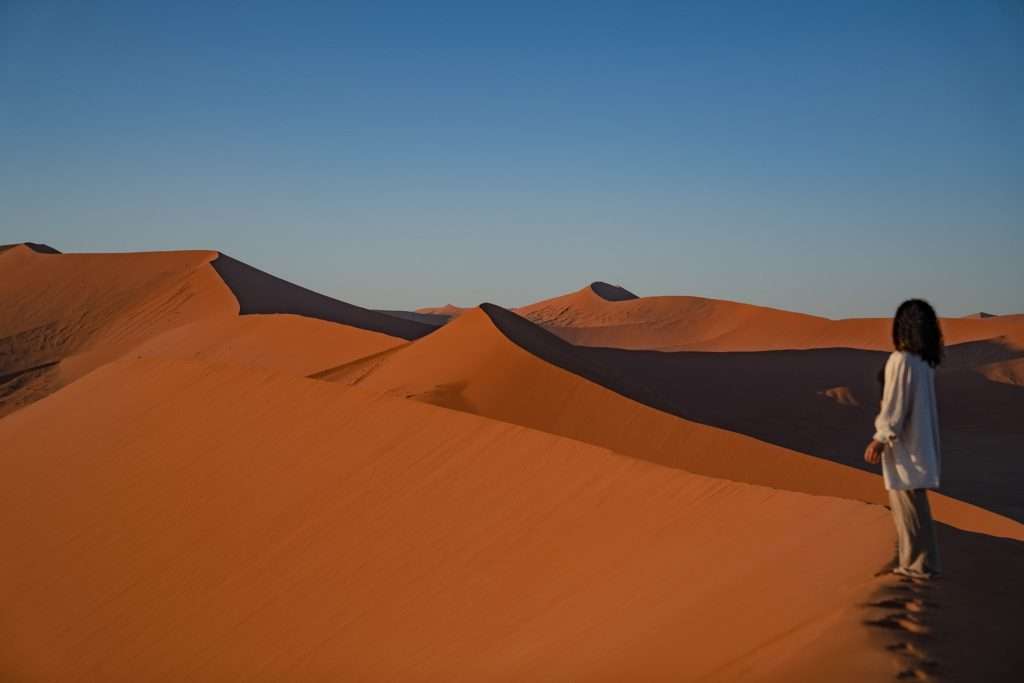 A person stands on the crest of a vast, rolling sand dune (Dune 45 In Namibia) in a desert, gazing out at the sweeping landscape under a clear blue sky. The orange sand dunes stretch far into the distance, creating a dramatic and serene vista, perfect for any 10-Day Namibia Itinerary.