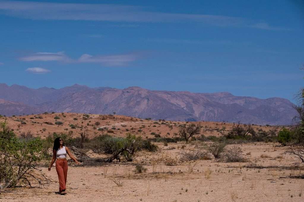 A person wearing a white top and rust-colored pants walks alone in a vast, arid desert landscape with sparse vegetation. Rocky mountains rise in the distance under a blue sky with a few scattered clouds, prompting the question: Is Namibia safe for solo female travelers exploring such serene yet isolated terrains?