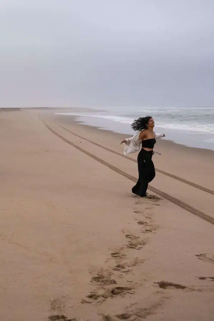A woman with curly hair, wearing black pants and a black top, runs joyfully along an empty sandy beach near the ocean—one of many serene moments you can experience on a 10-Day Namibia Itinerary. Tire and footprints mark the sand under a cloudy sky.