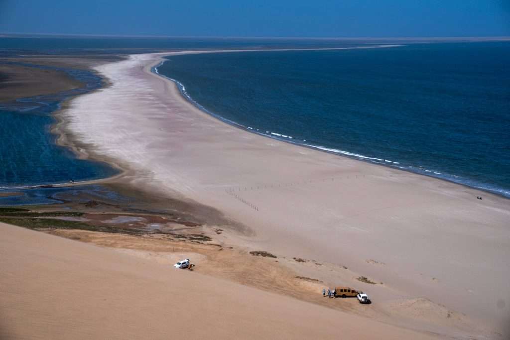 A scenic view of a sandy shoreline on the Skeleton Coast in Namibia curving along a deep blue ocean. The beach is bordered by a mix of light and dark sandy areas. Two vehicles and a trailer are parked on the sandy surface near the water's edge, with vast water stretching into the distance—a serene spot to ponder: Is Namibia safe for solo female travellers?