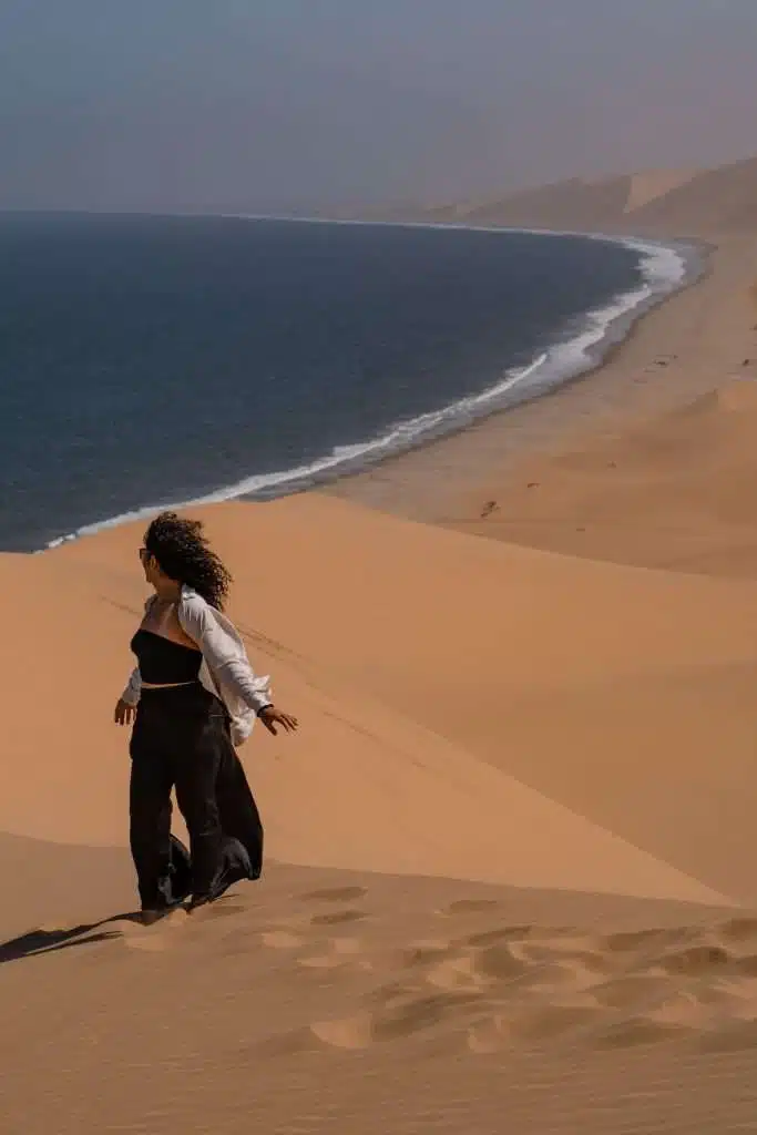 A person with curly hair walks down a sandy desert dune toward the ocean, waves meeting the shoreline under a hazy sky—an expansive, serene moment that sparks inspiration for a 10-Day Namibia Itinerary.