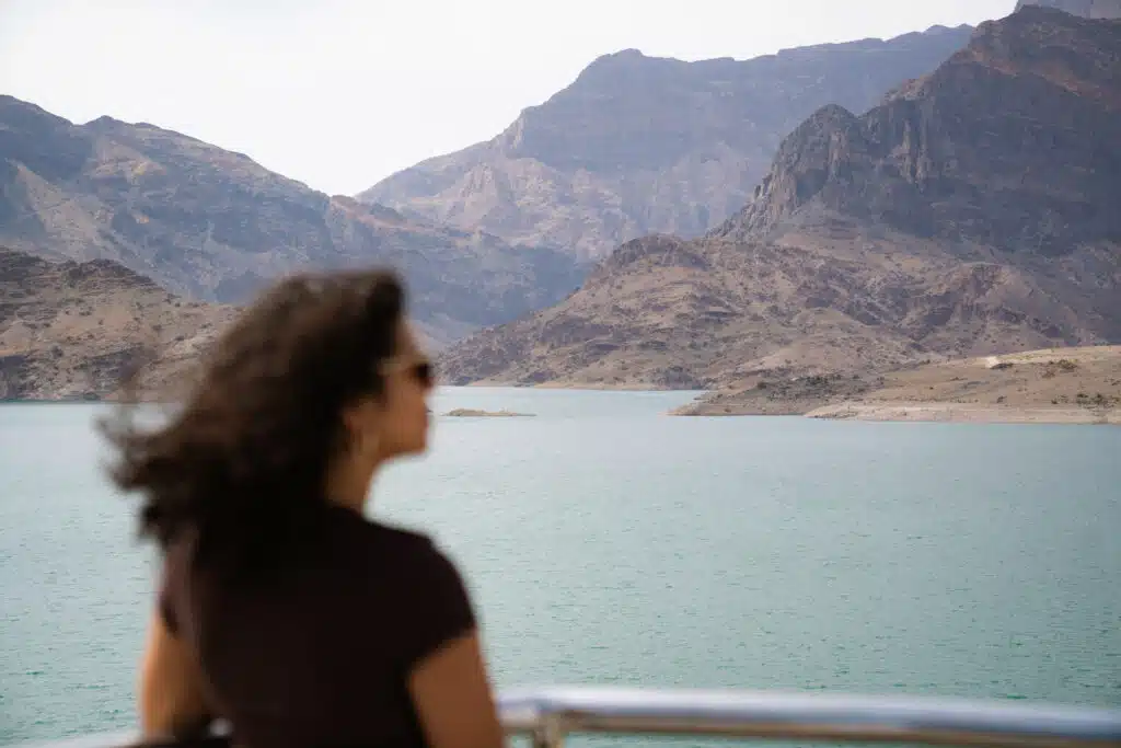 A person in their 30s, with curly hair and sunglasses, gazes at a serene body of water surrounded by rugged, brown mountains under a cloudy sky. The artistic blur of their face underscores the allure of backpacking through expansive landscapes.