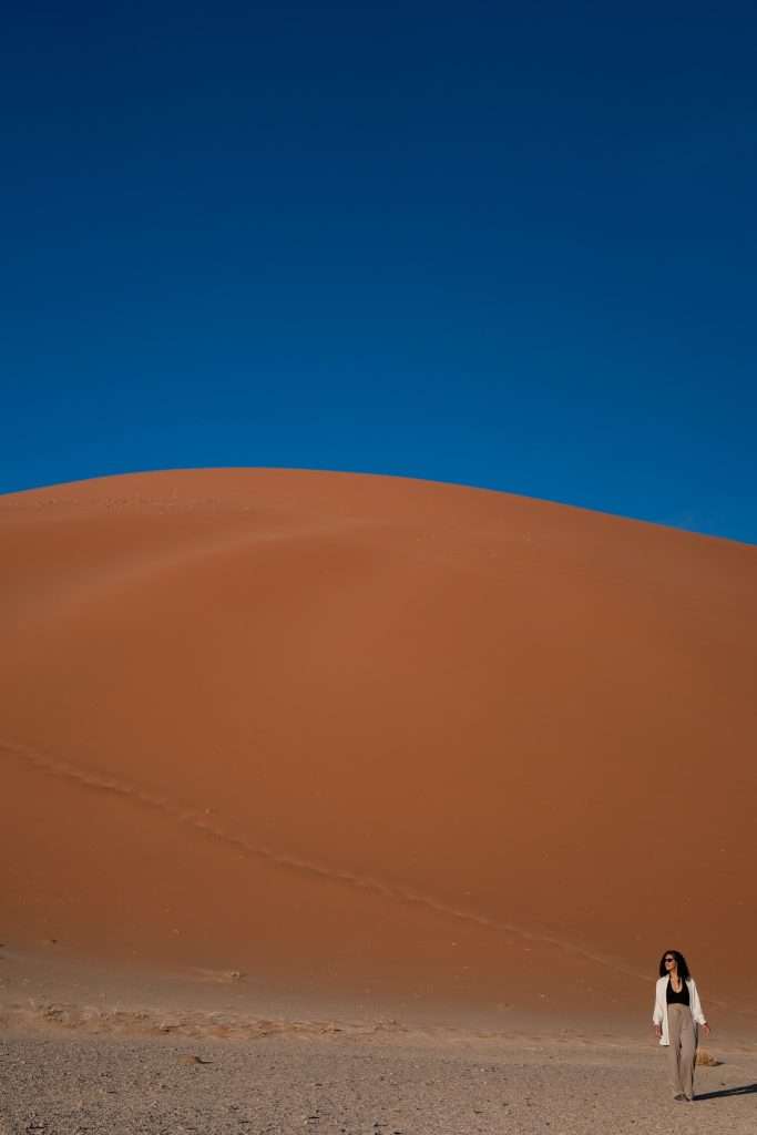 A person walks at the base of a vast, reddish  dune 45 in Namibia under a clear, deep blue sky. The dune's curved, smooth slope dominates the background, contrasting with the relatively flat, sandy terrain in the foreground. Amidst this natural beauty, one might ponder: Is Namibia safe for solo female travellers?