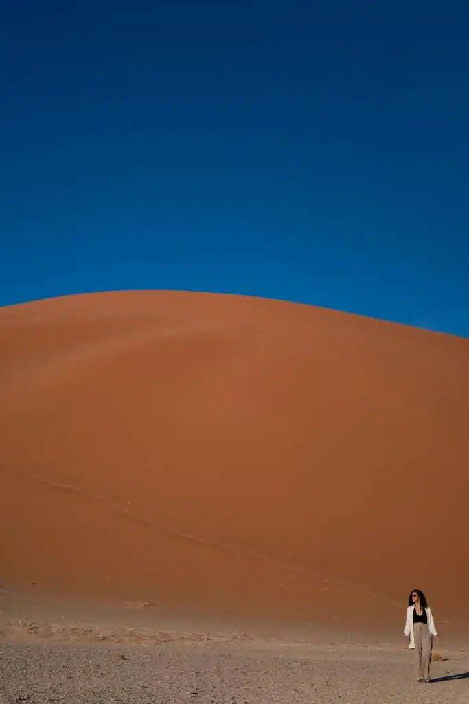 A person walks at the base of a vast, reddish dune 45 in Namibia under a clear, deep blue sky. The dune's curved, smooth slope dominates the background, contrasting with the relatively flat, sandy terrain in the foreground. Amidst this natural beauty, one might ponder: Is Namibia safe for solo female travellers?