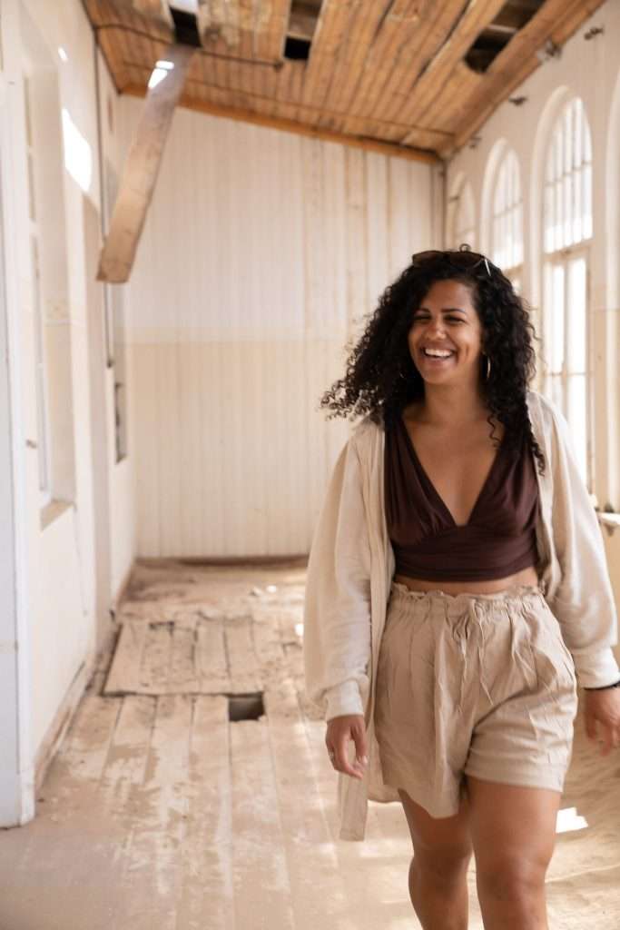 A person with curly hair and a big smile walks through a partially renovated room with wooden floors and a wooden ceiling. They are dressed casually in a brown top, light-colored shorts, and an open light-colored shirt. As natural light pours in through the windows, they ponder whether Namibia is safe for solo female travellers. - Kolmanskopp Namibia 