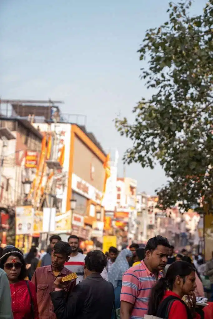 A busy street scene with many people walking in different directions, some wearing sunglasses and hats. Colorful shop signs suggest a lively area—raising questions like, "Is Varanasi Safe For Solo Female Travellers?" A tree is visible on the right side.