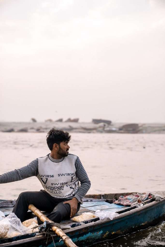 A man in a striped shirt rows a small wooden boat on a calm, hazy river. The sky is overcast, with rocks visible in the distance. Various items accompany him on his journey, reminiscent of essentials for a 4-week India itinerary.