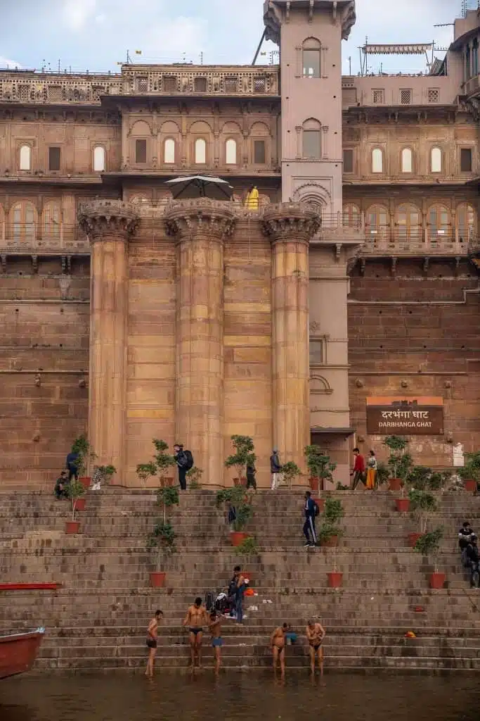 People stand and sit on wide stone steps leading to the river in front of the historic Darbhanga Ghat in Varanasi, India. Some bathe near the water while others gather on steps surrounded by potted plants, reflecting why many ask: Is Varanasi Safe For Solo Female Travellers?.