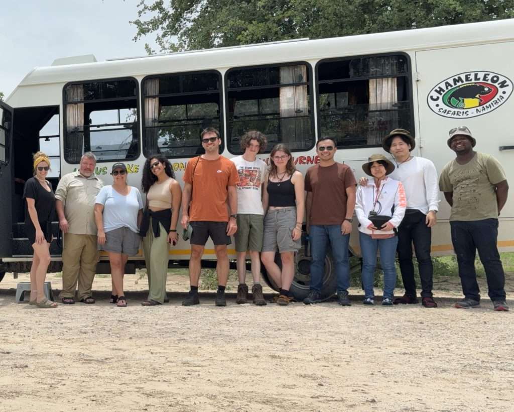 A diverse group of ten people stand in front of a bus labeled "Chameleon" on a gravel road. They appear to be on a tour, dressed casually for warm weather. Trees and part of a canopy can be seen in the background, suggesting an outdoor setting. One could wonder: Is Namibia safe for solo female travelers?