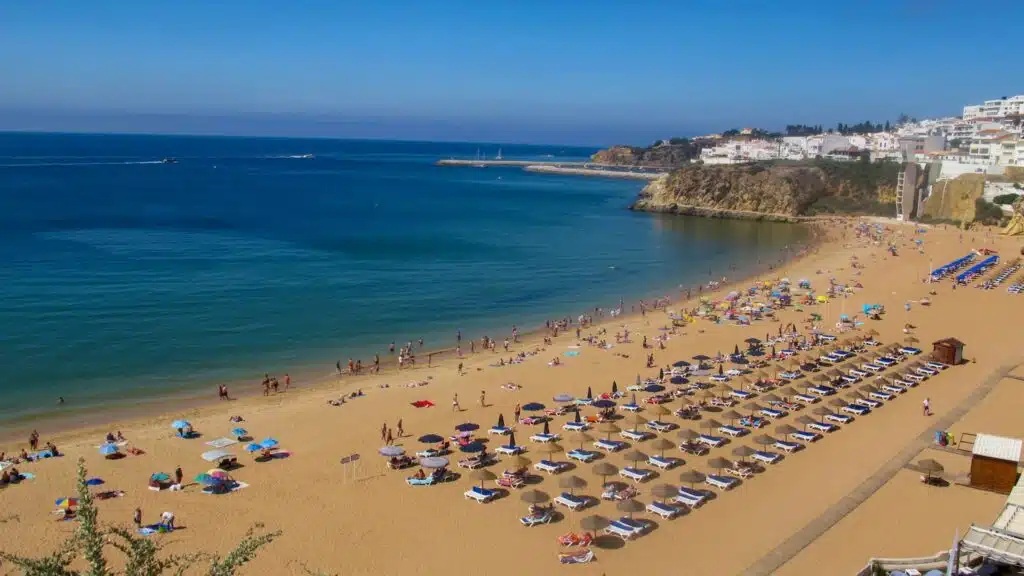 A sunny beach scene with rows of lounge chairs and umbrellas on golden sand, people relaxing and swimming, calm blue sea, and a coastal town on a rocky hill—ideal for those seeking the Best Boat Trips In The Algarve.