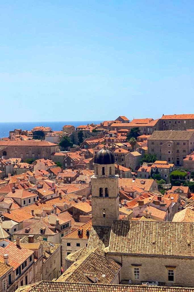 A scenic view of a historic Croatian town with beige stone buildings and orange-tiled roofs under a clear blue sky. A prominent bell tower stands in the foreground, perfect for your 7-day Croatia itinerary from Dubrovnik. The sea shimmers invitingly in the background.