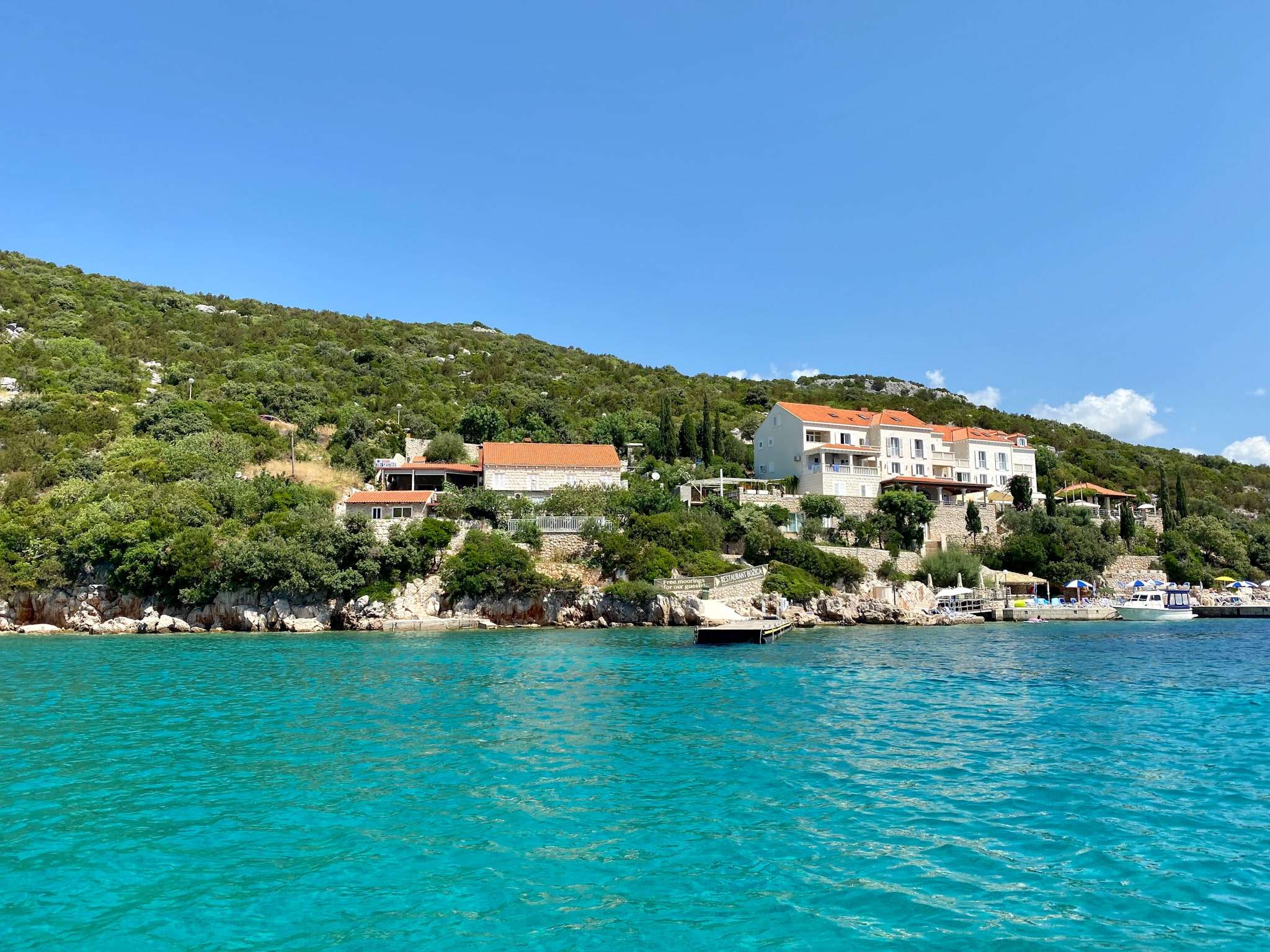 Scenic view of a coastal area with turquoise water in the foreground. Hills covered with greenery and several orange-roofed buildings are visible in the distance under a clear blue sky—a perfect scene for starting your 7-day Croatia itinerary from Dubrovnik.