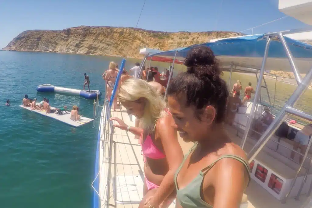 Two women in swimsuits stand on a boat deck, smiling and looking down at the water; people swim and relax on a floating platform nearby, with rocky cliffs in the background—capturing the vibe of the Best Boat Trips In The Algarve under a sunny sky.