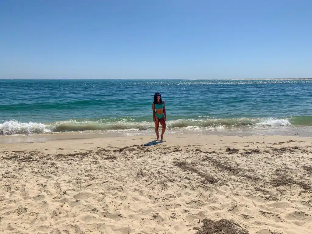 A woman in a turquoise bikini stands on a sandy beach near the shoreline, with gentle waves and a calm blue ocean under a clear, sunny sky—the perfect spot to discover the Best Boat Trips In The Algarve.