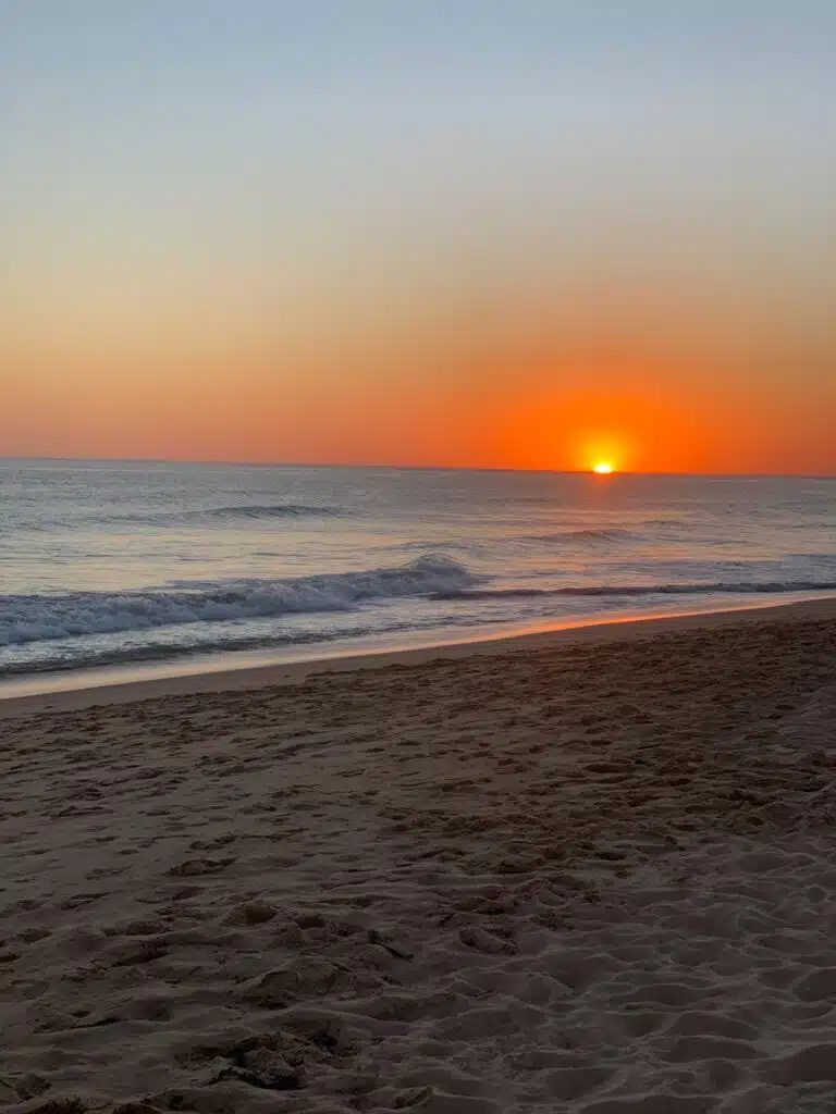 The sun sets over a calm ocean, casting an orange and yellow glow across the sky and gentle waves, with sandy beach in the foreground—an idyllic scene for enjoying the Best Boat Trips In The Algarve.