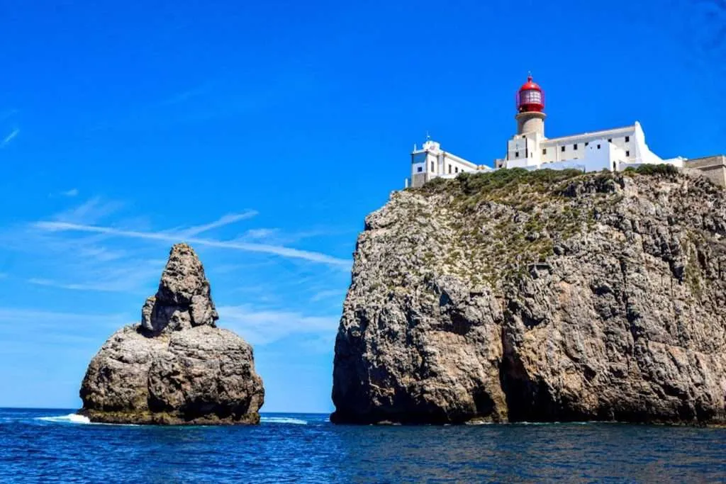 A lighthouse with a red dome sits on the edge of a rocky cliff by the sea, under a clear blue sky. A large rock formation rises from the water nearby, offering one of the best coastal sights for boat trips in the Algarve.