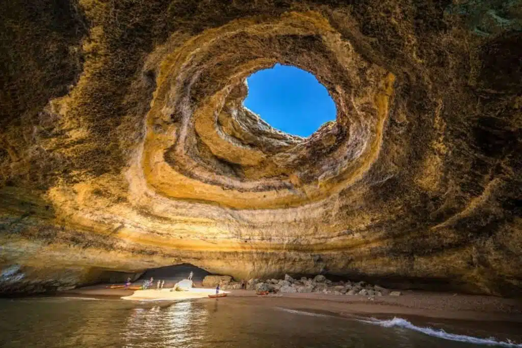 A large, natural cave with a circular opening at the top allows sunlight to illuminate the sandy interior. The cave features layered rock formations, perfect for those exploring by foot or enjoying the best boat trips in the Algarve, as they venture into shallow waters at the entrance.
