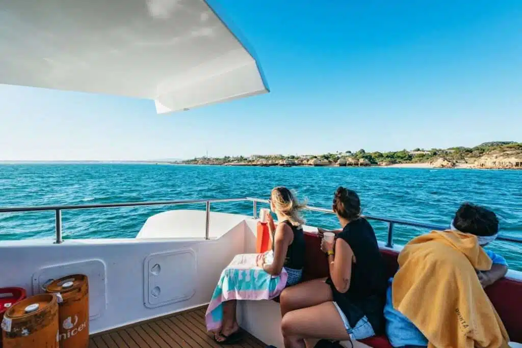 People sitting on a boat under a sunny sky, with two women looking toward the coastline. One person is wrapped in a yellow towel. Lush greenery borders the ocean, and blue water surrounds the vessel, epitomizing one of the best boat trips in The Algarve.