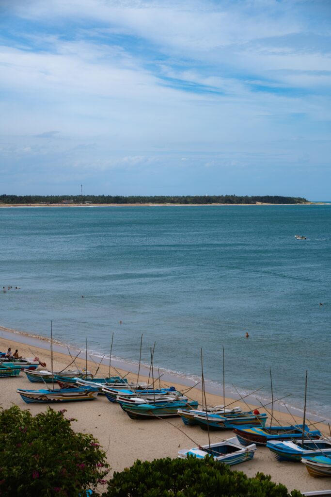 A row of colorful wooden fishing boats is lined up on a sandy beach in Arugam Bay beside calm blue water, with a distant shoreline and cloudy sky beyond—one of the top things to do in Arugam Bay as people swim in the sea nearby.