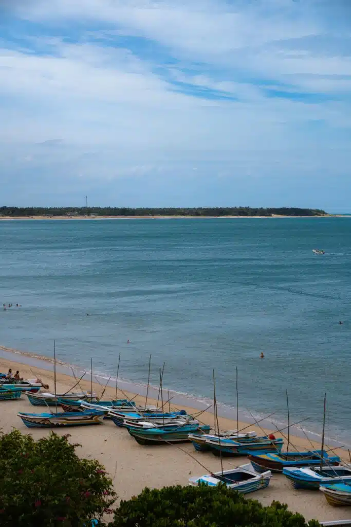 A row of colorful wooden fishing boats is lined up on a sandy beach in Arugam Bay beside calm blue water, with a distant shoreline and cloudy sky beyond—one of the top things to do in Arugam Bay as people swim in the sea nearby.