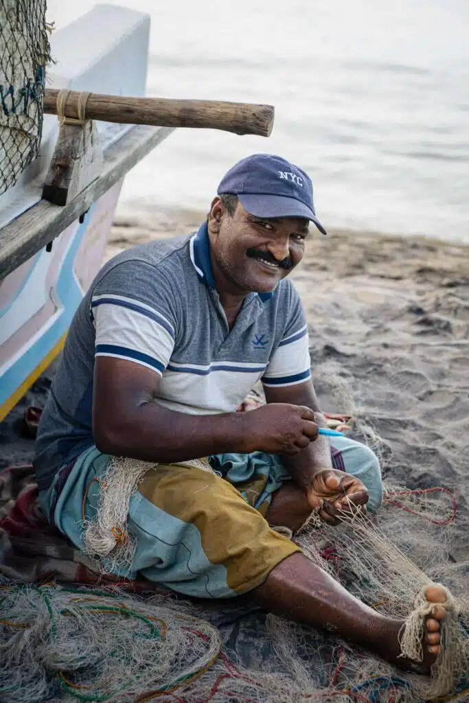 A smiling man wearing a cap and striped polo shirt sits on the sand by the shore, mending fishing nets next to a boat—capturing authentic local life among the many Things To Do In Arugam Bay.
