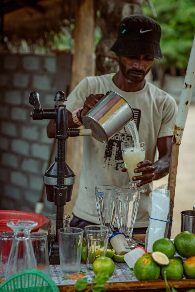A man in a black hat and beige shirt pours fresh juice from a metal jug into a blender at an outdoor juice stand—one of the vibrant things to do in Arugam Bay, with limes, glasses, and a manual juicer on the counter.