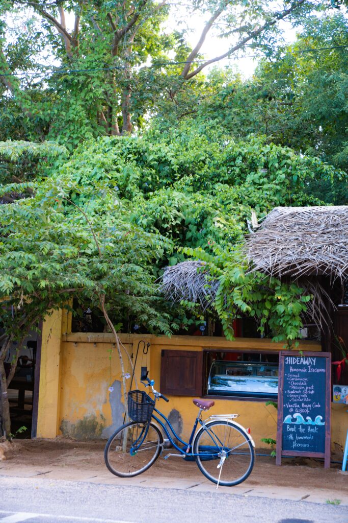 A blue bicycle with a basket is parked in front of a yellow wall and a rustic café with thatched roofs—a perfect scene to add to your list of Things To Do In Arugam Bay. A chalkboard menu stands nearby, framed by lush green trees.