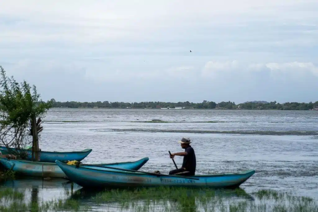 A person wearing a hat sits in a blue canoe near the shore of a calm, wide river—one of the peaceful things to do in Arugam Bay—with grassy banks, trees, and distant land visible under a cloudy sky.