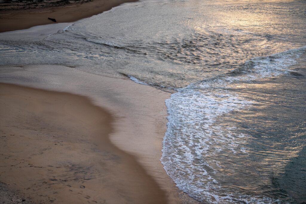 Aerial view of gentle ocean waves washing onto a sandy beach at sunset, with soft light reflecting on the water—one of the many picturesque things to do in Arugam Bay, where smooth sand lines the scenic shoreline.