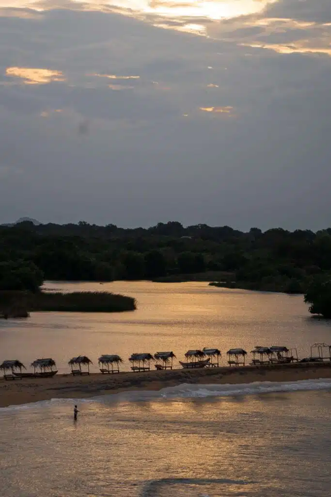A tranquil beach at sunset with several small thatched huts lined up on the sand, calm water in the foreground, and lush greenery in the background—one of the serene things to do in Arugam Bay as a person stands at the water’s edge.