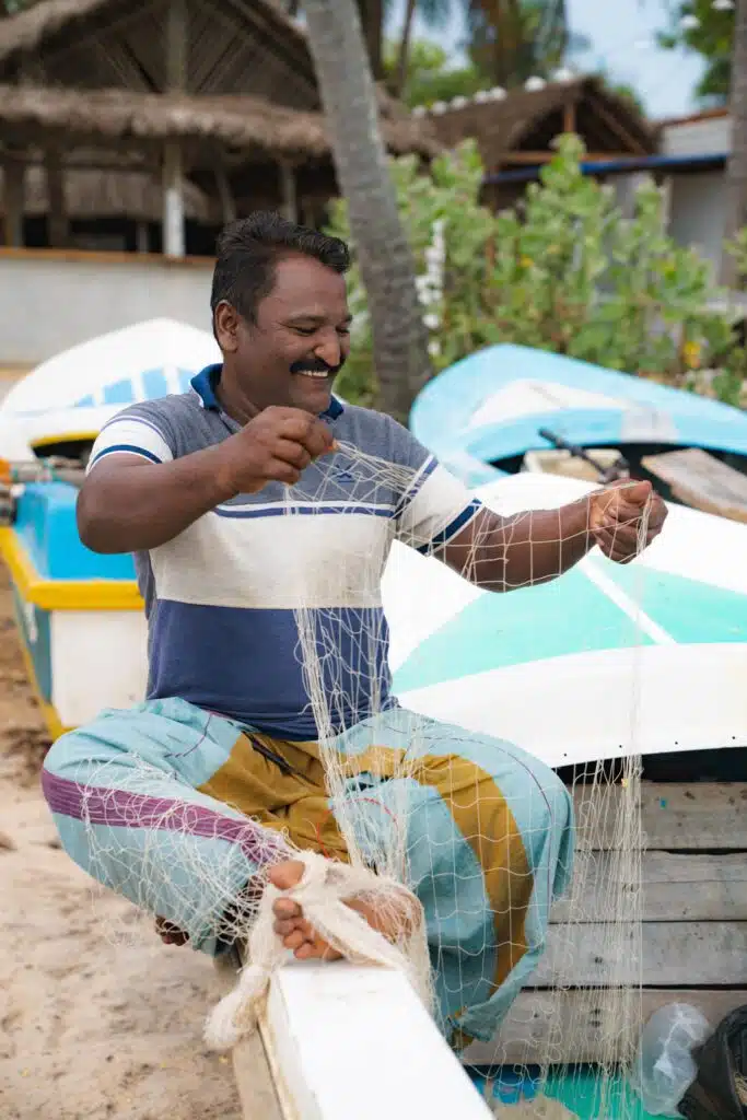 A man sits on a dock by boats, smiling as he repairs a fishing net with his hands and feet. Tropical plants and a thatched-roof building are visible, capturing the laid-back vibe of Things To Do In Arugam Bay.